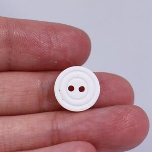May include: A small, white button with two holes and a concentric ring design. The button is round and appears to be made of plastic. It is held in a person's hand against a plain white background. Suitable for sewing and craft projects.