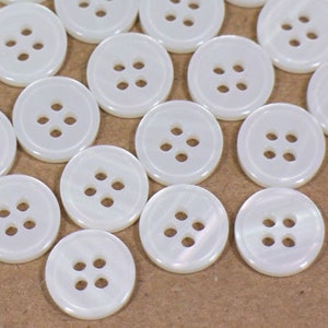 以下が含まれることがあります： 白くて丸いボタンのコレクションで、それぞれに4つの穴があります。ボタンは茶色の表面に散らばっています。これらの縫製用品は、さまざまな工芸品やアパレルプロジェクトに最適です。