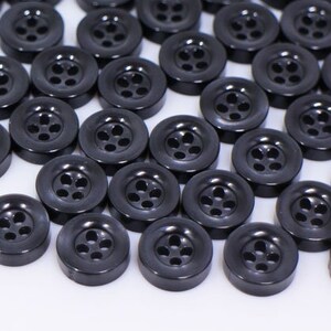 Peut inclure: Un grand groupe de boutons en plastique noir avec cinq trous chacun. Les boutons sont disposés en un tas lâche sur une surface blanche.