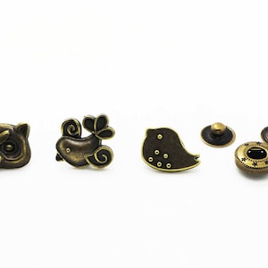 May include: Four metal buttons with different animal designs. The buttons are in antique gold and feature an owl, a bird with a leaf, a bird with dots, and a round button with a star design.