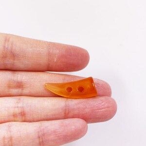 May include: A close-up of a small, orange, plastic button shaped like a horn with three holes.