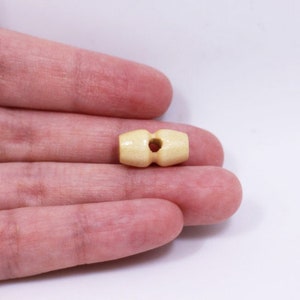 May include: A close-up of a wooden toggle button in a light beige colour. The button has a hole in the centre and is designed for use in clothing or crafts.
