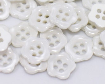 Könnte beinhalten: Eine Sammlung weißer Plastikknöpfe mit jeweils vier Löchern. Die Knöpfe sind blumenförmig mit abgerundeten Kanten.