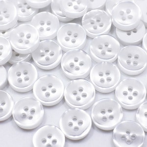以下が含まれることがあります： 白いプラスチック製のボタンが4つの穴が開いていて、白い表面にランダムに配置されています。