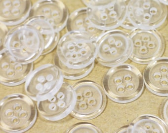 May include: A large pile of clear plastic buttons with four holes each. The buttons are scattered on a brown surface.