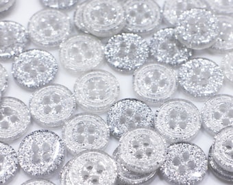 May include: A large group of silver glitter buttons with four holes each. The buttons are scattered on a white surface.