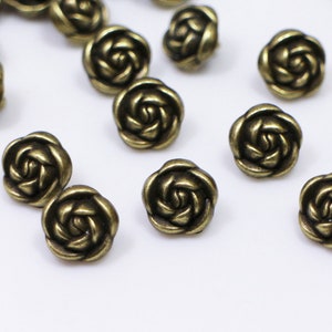 May include: A collection of antique gold-toned metal rose-shaped buttons. The buttons have a raised, detailed design and are scattered on a white surface.