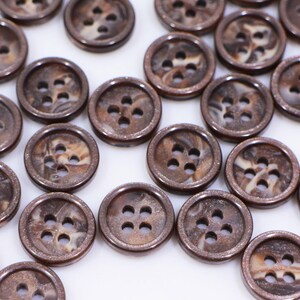 May include: A collection of brown and white patterned buttons with four holes each. The buttons are scattered on a white surface.
