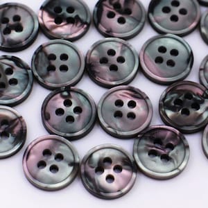 May include: A collection of black and iridescent buttons with four holes each. The buttons have a marbled pattern and are arranged in a random pattern on a white surface.
