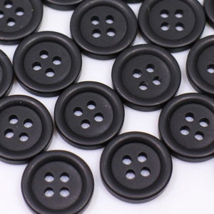 May include: A large group of black plastic buttons with four holes each. The buttons are scattered on a white surface.