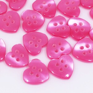 Könnte beinhalten: Eine Gruppe von rosa herzförmigen Knöpfen mit jeweils vier Löchern. Die Knöpfe sind auf einer weißen Oberfläche verstreut.