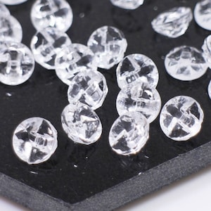 Könnte beinhalten: Nahaufnahme eines Stapels durchsichtiger Plastikknöpfe auf einer schwarzen sechseckigen Oberfläche. Die Knöpfe sind über die Oberfläche verteilt und einige fallen über den Rand.