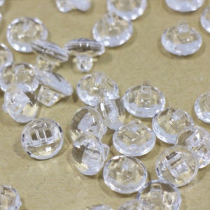 May include: A large group of clear plastic buttons with a faceted design. The buttons are scattered on a brown surface.