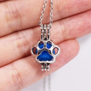 May include: A silver charm necklace with a blue paw print charm. The charm is open and can be used to hold a small object, such as a photo or a piece of hair.