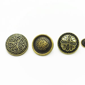 May include: Five antique-style metal buttons with intricate gold designs on a black background. The buttons are round and have a raised edge. Two of the buttons have a floral design, one has a geometric design, and two are smaller and have a star-shaped design.