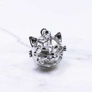 Könnte beinhalten: Eine silberne Halskette mit einem Katzen-Anhänger und einer zarten Kette. Der Anhänger ist eine hohle Kugel mit einem Katzenkopf-Design.