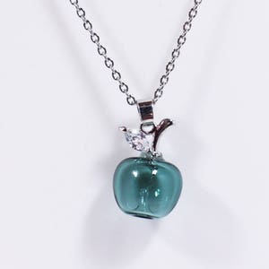 Op de afbeelding: Zilverkleurige ketting met een hanger in de vorm van een teal appel. De hanger heeft een zilverkleurige steel en een kleine vogelachtige versiering met heldere kristallen. De ketting is delicaat, tegen een witte achtergrond.