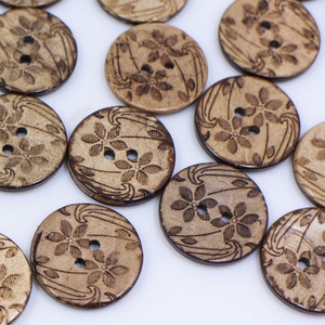 May include: A collection of brown coconut shell buttons with a floral design. The buttons have two holes and are arranged in a scattered pattern on a white background.
