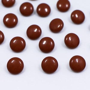 Könnte beinhalten: Eine Sammlung brauner Plastikknöpfe, verstreut auf einer weißen Oberfläche. Die Knöpfe sind rund und haben eine glatte, glänzende Oberfläche.