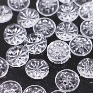 May include: A collection of clear plastic buttons with a decorative, starburst-like design. The buttons are arranged on a black surface.