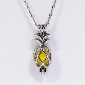 Könnte beinhalten: Eine silberne Halskette mit einem ananasförmigen Anhänger, in dem sich eine gelbe Glaskugel befindet. Der Anhänger ist mit einer Ananasspitze und einem käfigartigen Design verziert.