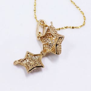 May include: A gold-colored star-shaped pendant necklace. The pendant opens to reveal a hollow interior. The necklace chain is delicate and also gold-colored. The pendant has a detailed, openwork design. The necklace is displayed against a white background.