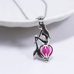 Peut inclure: Un collier en argent avec un pendentif en forme de sirène contenant une perle rose. La sirène est assise sur une coquille et a de longs cheveux flottants.