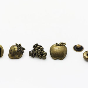 May include: A set of six antique gold-toned metal buttons featuring fruit designs. The buttons include a banana, strawberry, grapes, apple, and two round buttons with a star design.