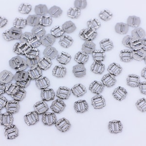 May include: A large group of silver metal slider buckles with a decorative design. The buckles are scattered on a white surface.