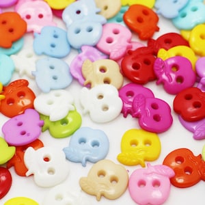 May include: A collection of colorful, apple-shaped buttons in various shades, including red, pink, yellow, blue, and green. Each button has two holes for sewing. The buttons are scattered on a white surface.