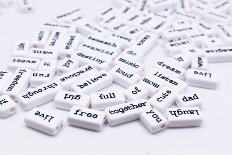 May include: White rectangular beads with black lettering spelling out words like "love", "dream", "music", "joy", "believe", "together", "free", "full", "cute", "dad", "listen", "wow", "laugh", and "run".
