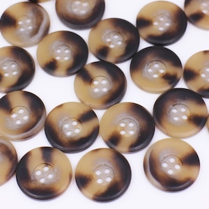 Könnte beinhalten: Eine Sammlung von braunen und schwarzen gemusterten Knöpfen mit jeweils vier Löchern. Die Knöpfe sind aus einem glatten, glänzenden Material gefertigt und haben einen leicht erhöhten Rand.
