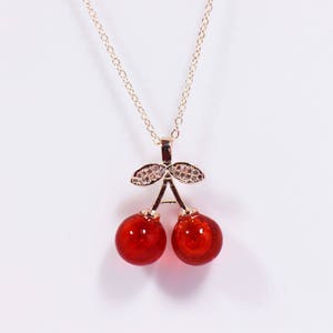 Op de afbeelding: Een gouden ketting met een rode kersen hanger. De hanger heeft twee rode kersen met gouden bladeren en kleine heldere stenen.