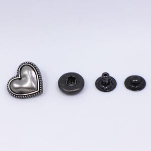 May include: A silver heart-shaped button with a beaded border and a black metal snap fastener set. The set includes a stud, socket, and cap.