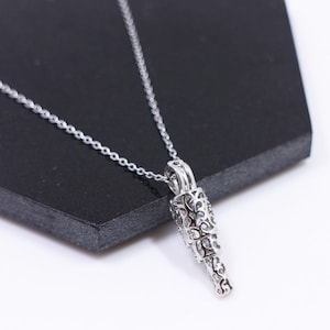 以下が含まれることがあります： 装飾的なデザインが施された、細部までこだわったペンダントがついたシルバーネックレス。ペンダントは、長い薄い円筒形をしています。