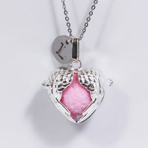 Peut inclure: Collier en argent avec un pendentif en forme de cœur orné d'ailes d'ange et d'un intérieur rose. Le pendentif est attaché à une chaîne en argent avec un petit charme en argent en forme de disque gravé de la lettre "L".