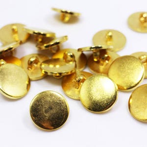 Könnte beinhalten: Eine Gruppe von 14 goldfarbenen Metallknöpfen mit einem Stiel auf der Rückseite. Die Knöpfe sind rund und haben eine glatte, glänzende Oberfläche.