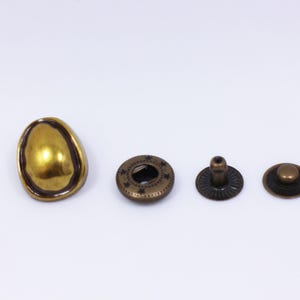 May include: A collection of four antique brass-colored metal fasteners. The set includes a large, irregular-shaped button, a decorative snap with star-shaped cutouts, a snap post, and a small button.