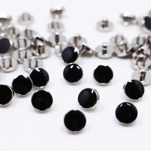 以下が含まれることがあります： 黒と銀の丸いボタンのグループ。ボタンは平らな円形のトップで、黒の中央と銀の縁があります。ボタンは、布に取り付けるための小さな銀のポストを背面に備えています。