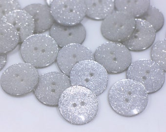 May include: A collection of silver glitter buttons with two holes each. The buttons are round and have a shiny, sparkly finish.