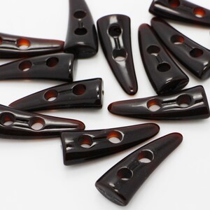May include: A group of brown plastic horn-shaped buttons with two holes each. The buttons are arranged in a random pattern on a white surface.