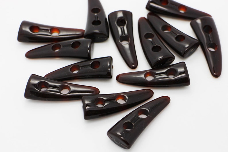 Peut inclure: Un groupe de boutons en plastique brun en forme de corne avec deux trous chacun. Les boutons sont dispos&eacute;s de mani&egrave;re al&eacute;atoire sur une surface blanche.