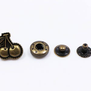 May include: A set of four antique gold-toned metal snap fasteners. One fastener features a cherry design, while the other three are round with a raised center.