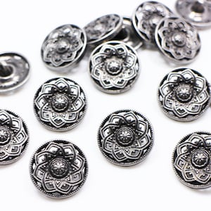 Könnte beinhalten: Eine Sammlung silberfarbener Metallknöpfe mit filigranen Blumendesigns. Die Knöpfe sind rund und haben eine erhabene Mitte.