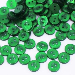 May include: A collection of shiny, emerald green buttons with two holes each. The buttons are round and have a slightly raised edge, scattered on a white surface. These buttons are ideal for sewing and craft projects.