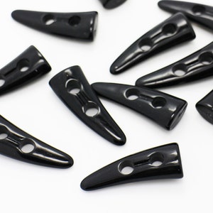 May include: A group of black plastic horn-shaped buttons with two holes each. The buttons are scattered on a white surface.