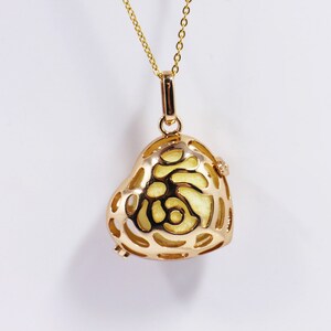 Könnte beinhalten: Eine goldene Halskette mit einem herzförmigen Anhänger mit einem filigranen, komplizierten Design. Der Anhänger ist hohl und hat ein hellgelbes Inneres. Die Halskette besteht aus einer feinen Kette.