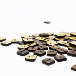 K&ouml;nnte beinhalten: Eine Sammlung von braunen und beigefarbenen Kokosnuss-Kn&ouml;pfen mit jeweils zwei L&ouml;chern. Die Kn&ouml;pfe sind auf einer wei&szlig;en Oberfl&auml;che verstreut.