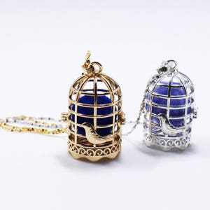 May include: Two birdcage necklaces, one gold and one silver, with blue velvet interiors. The necklaces are displayed on a white background.
