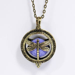 以下が含まれることがあります： 紫色のトンボが閉じ込められた丸いペンダントが付いたゴールドトーンのネックレス。ペンダントは、細部までこだわったトンボのデザインが施された複雑なデザインです。